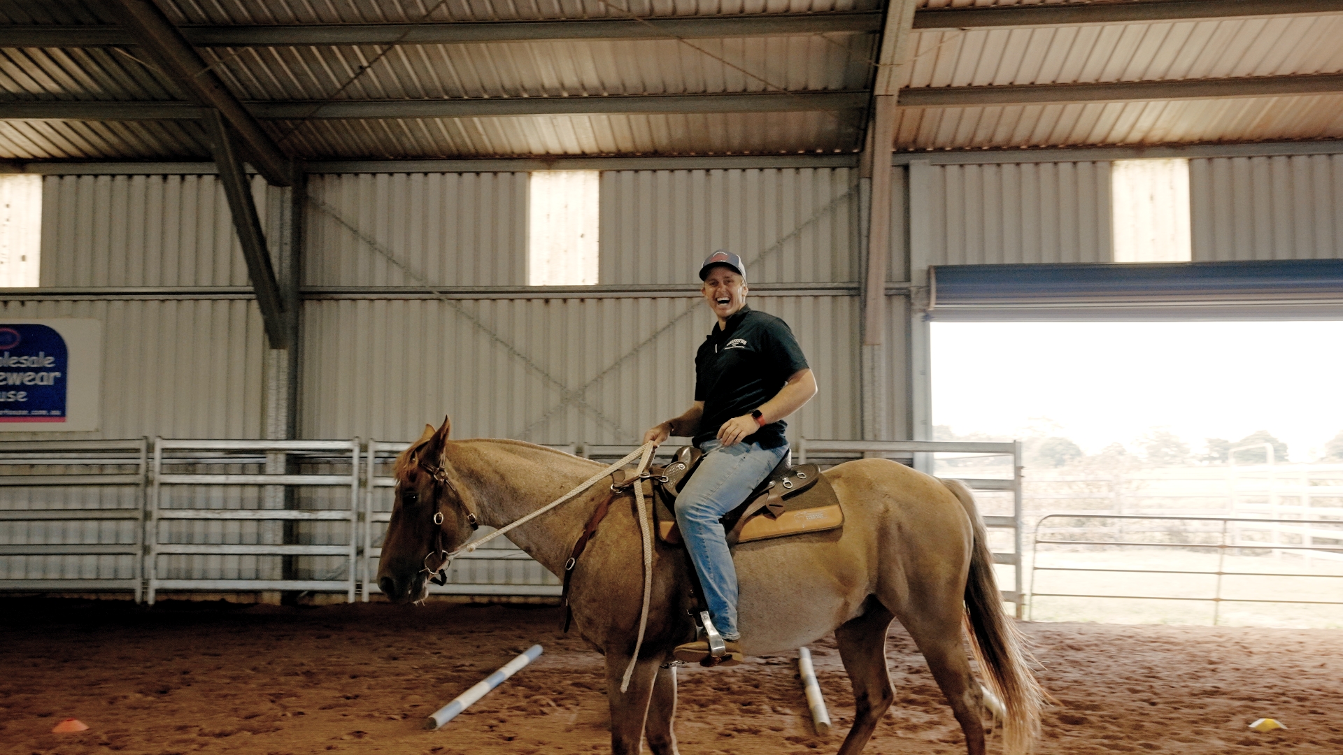 A smiling man wearing a cap and black shirt rides a light brown horse inside an indoor riding arena.