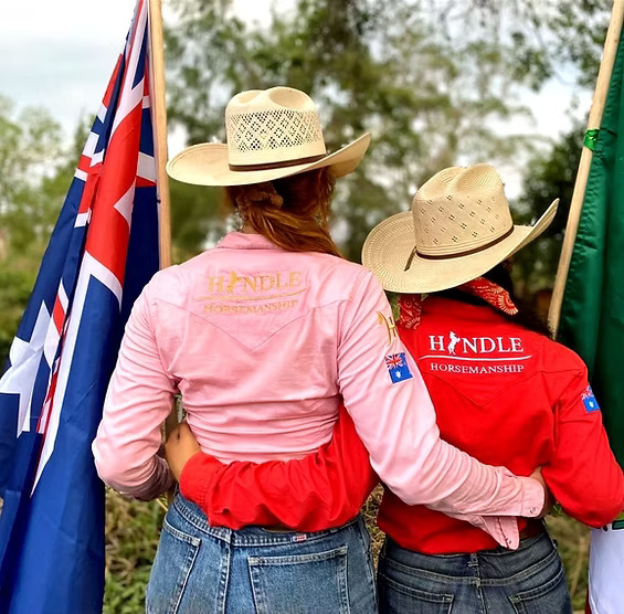 Two people in cowboy hats and denim jeans hugging, each holding a flag, wearing shirts labeled 'Hindle Horsemanship.'