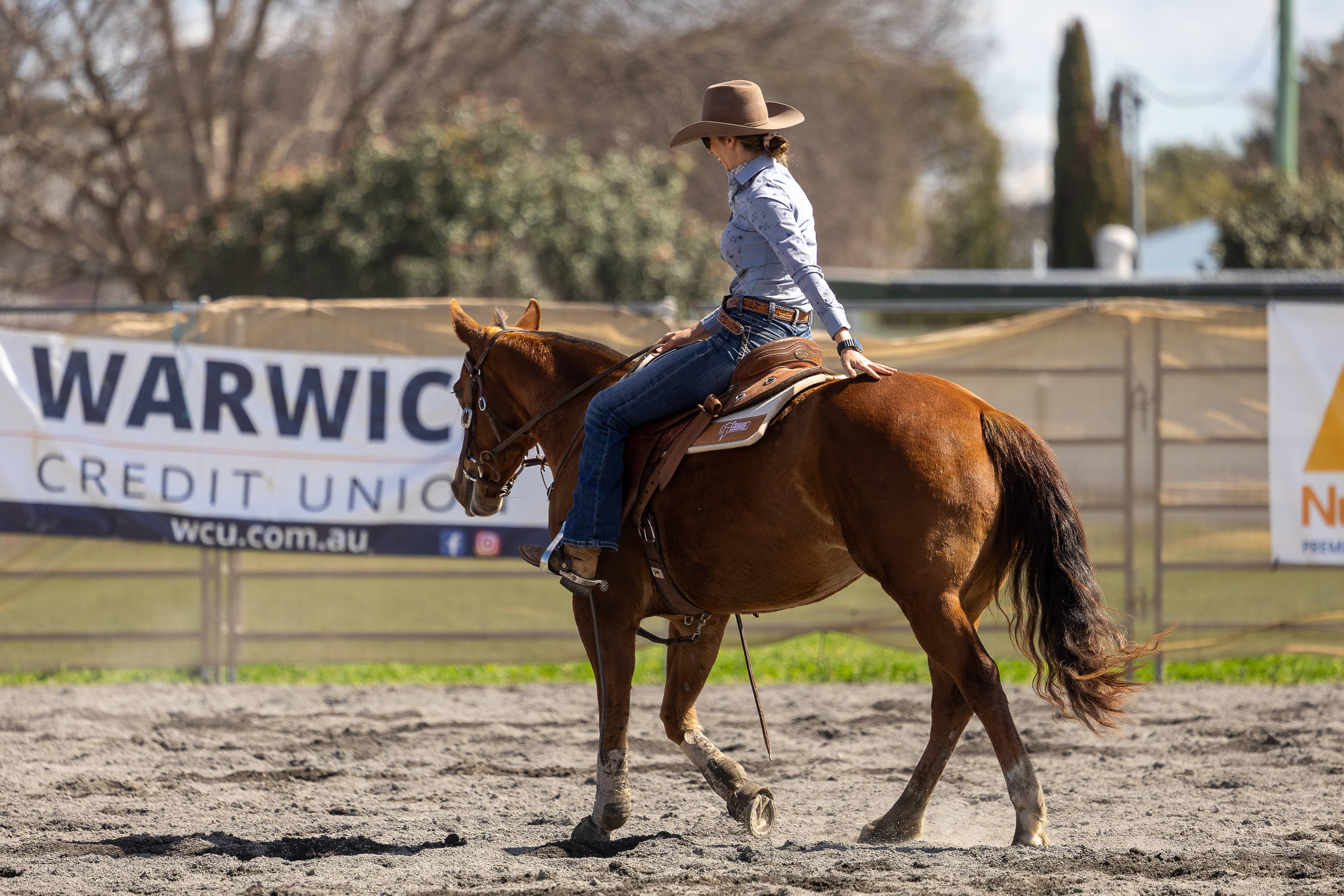 Woman wearing a cowboy hat riding a brown horse in an outdoor arena with a Warwick Credit Union banner in the background.