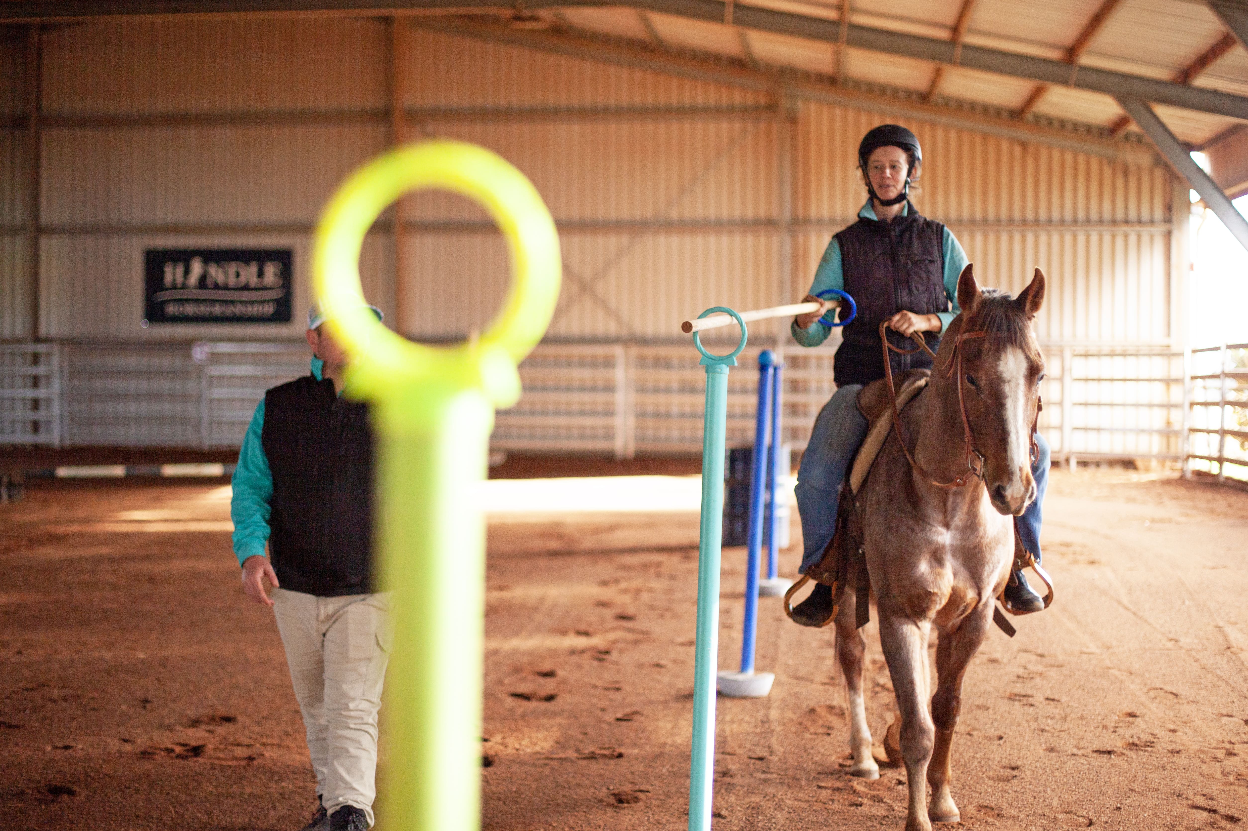 Woman wearing helmet riding a horse indoors while holding a long pole through colored ring stands, guided by a man walking beside them.