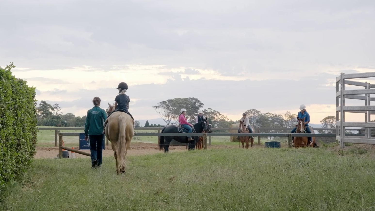 Person walking beside a child on a horse with three other riders sitting on horses in a fenced grassy area under a cloudy sky.
