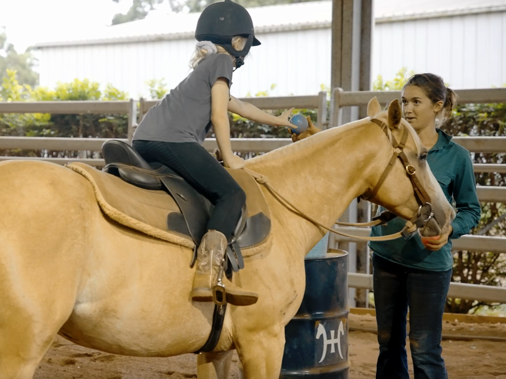 A child wearing a helmet on a horse reaching to hand a blue ball to a standing woman holding a red ball in a fenced area.