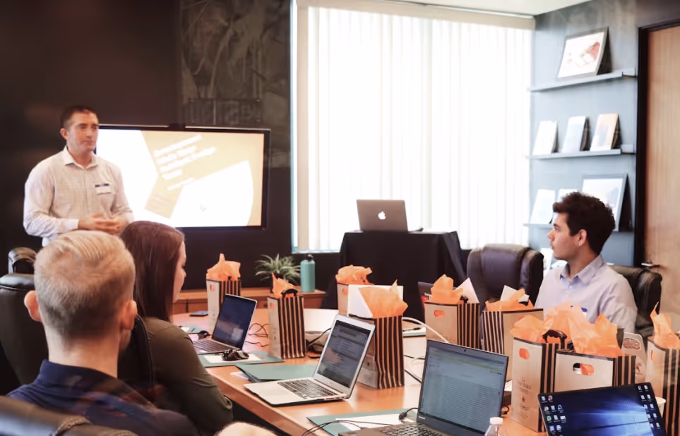 A man in a white shirt giving a presentation to a group of four people seated around a conference table with laptops and gift bags.
