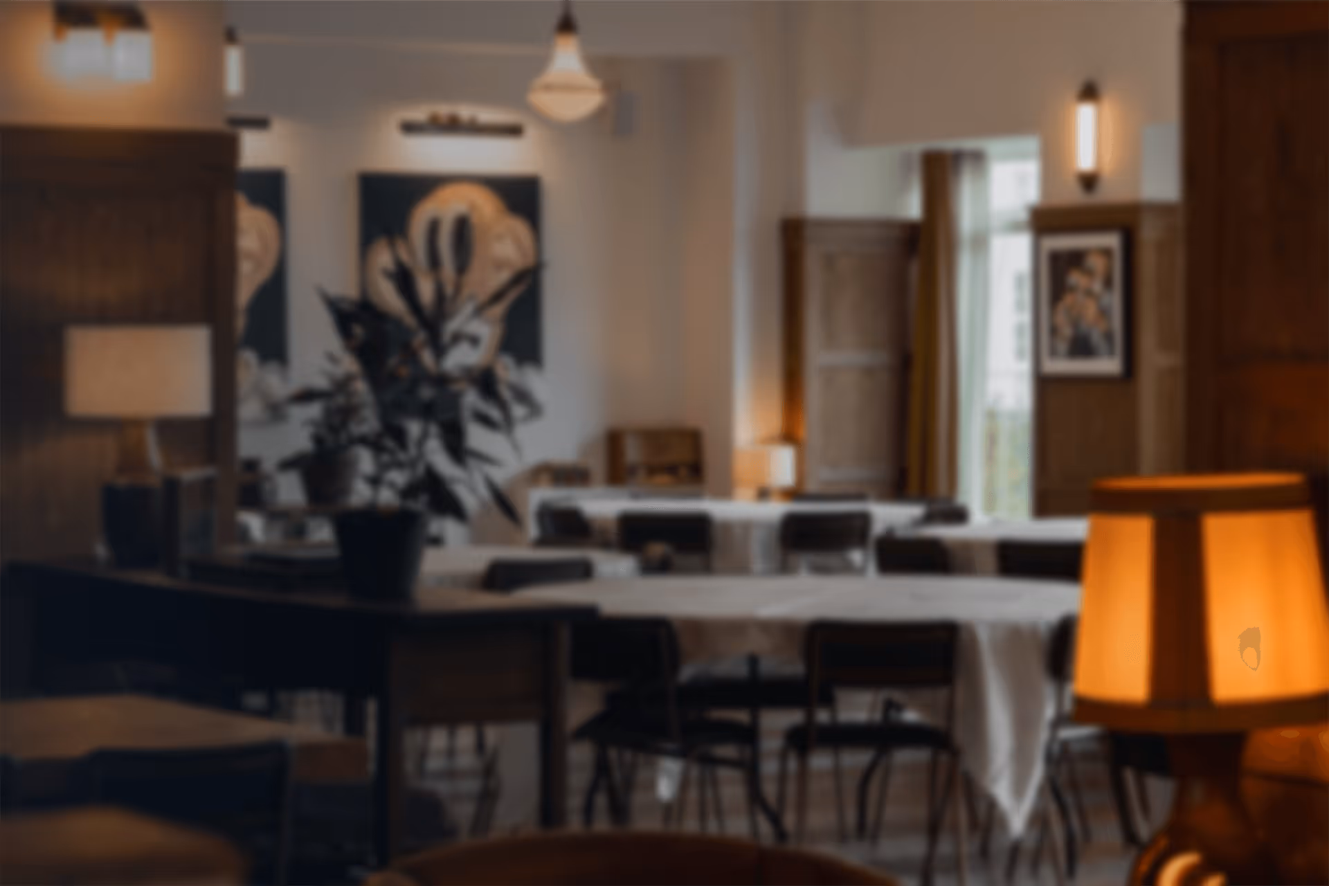 Blurry image of a warmly lit dining area with tables, chairs, plants, and framed artwork on the walls.
