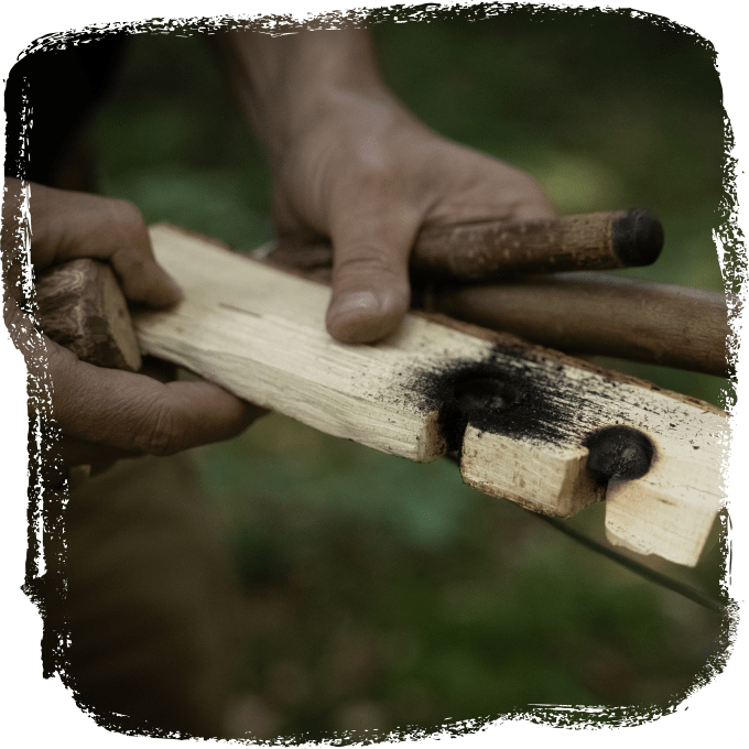 Zwei Hände halten zwei Holzstücke, eines davon hat verkohlte Stellen, während ein Holzstab daran gerieben wird.