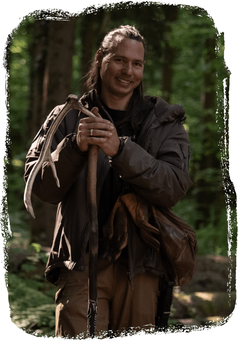 Gion Saluz mit langem Haar im Wald hält ein Geweih und lehnt sich an einen Stock, trägt eine Jacke und braune Hosen.