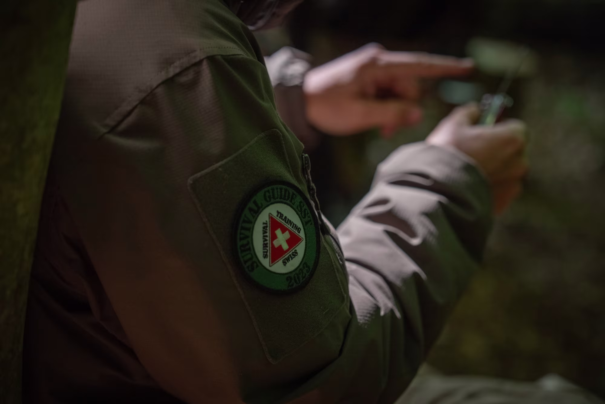Nahaufnahme eines Armes in olivgrüner Jacke mit einem Swiss Survival Guide Trainingsabzeichen auf dem Ärmel, die Person zeigt mit der Hand auf ein Werkzeug.