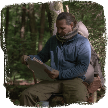 Mann mit Rucksack sitzt im Wald auf einem Baumstumpf und blickt auf eine Karte.