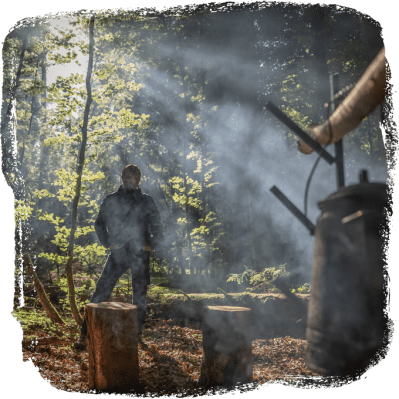 Person steht im Wald vor drei Baumstümpfen, während Rauch durch Sonnenlicht schimmert und eine Hand eine Kaffeekanne hält.