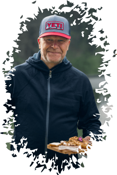 Mann mit grauer YETI-Kappe und schwarzer Jacke hält ein Holzbrett mit verschiedenen Lebensmitteln.