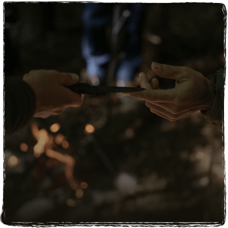 Zwei Personen, die im Freien bei einem Lagerfeuer ein mündliches Dokument oder Gerät überreichen.