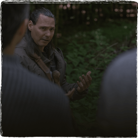 Mann mit langem Haar spricht im Wald mit zwei weiteren Personen.