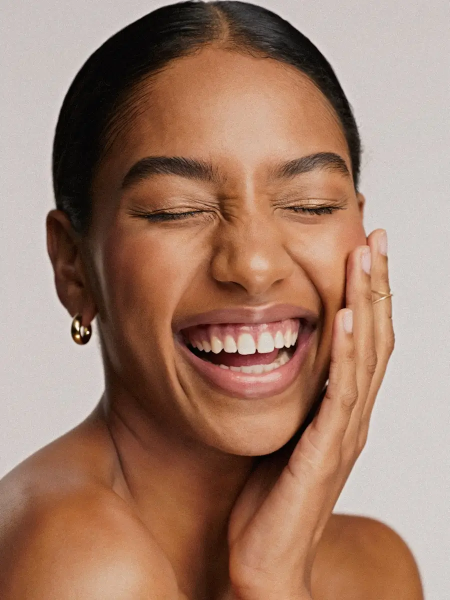 Close-up portrait of a radiant woman with eyes closed and a wide smile, touching her face with one hand.