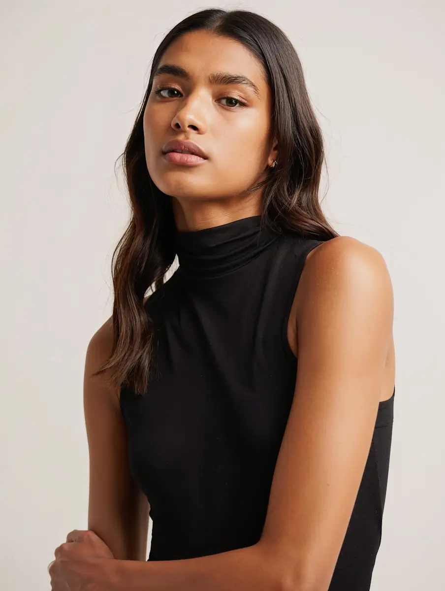 Portrait of a woman with long dark hair wearing a sleeveless black turtleneck against a light background.