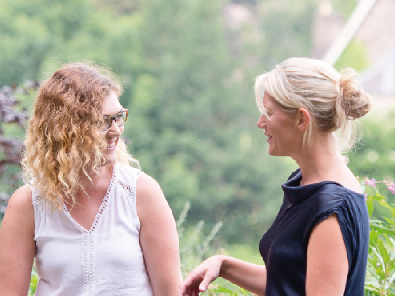 Two women smiling and engaging in conversation outdoors against a soft, blurred green background of trees and foliage.