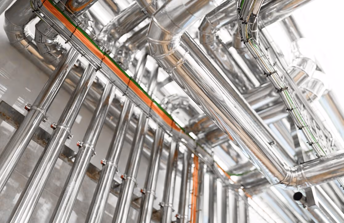 Network of shiny stainless steel industrial pipes and conduits in a mechanical room.