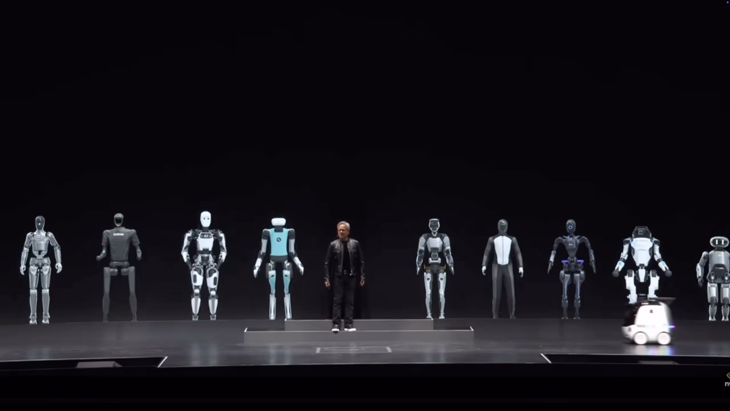 A man standing on a stage surrounded by various humanoid robots in different designs and colors, with one small wheeled robot moving in front.