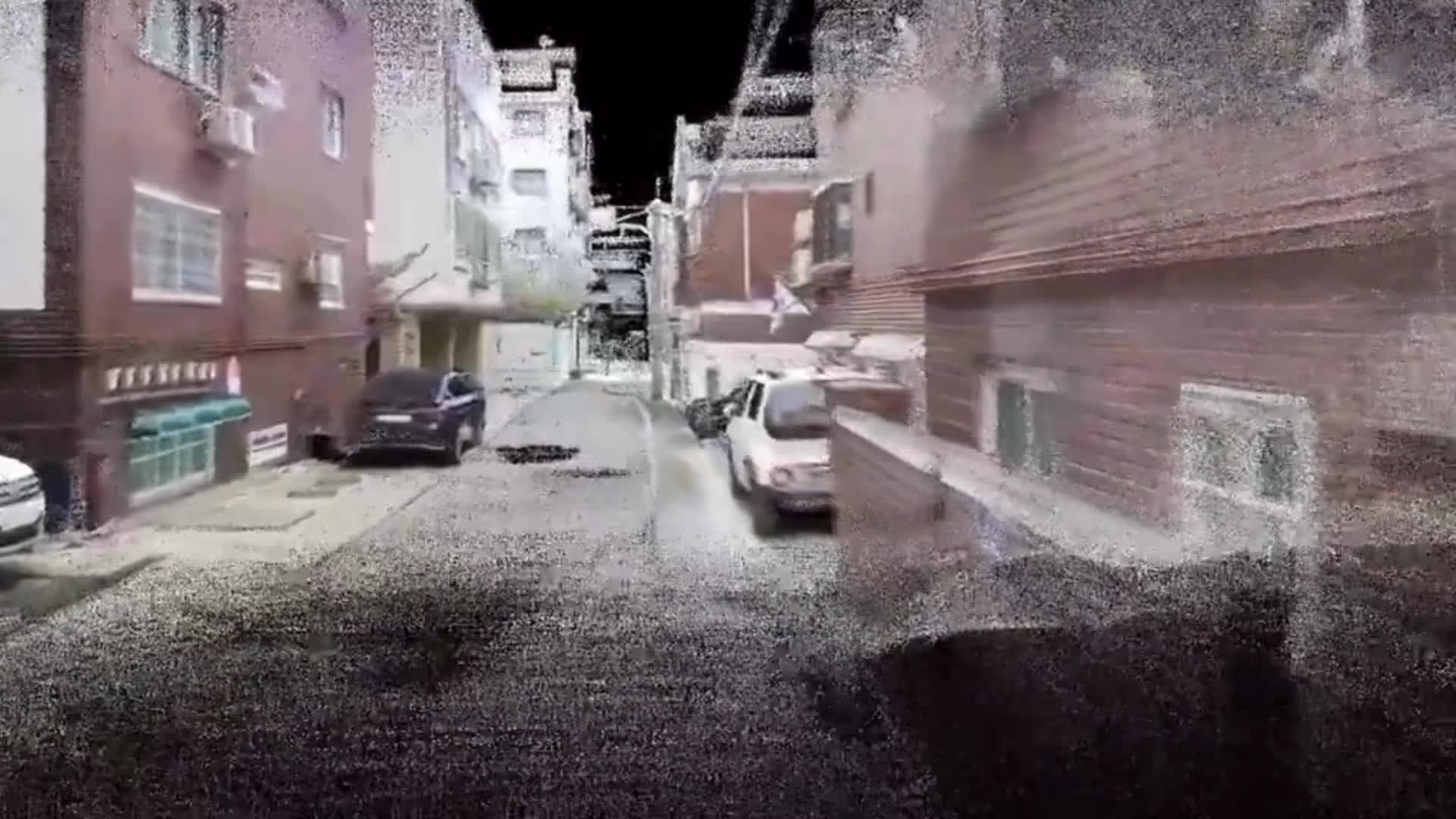 Grainy, pixelated street view with parked cars and buildings on both sides under a black sky.