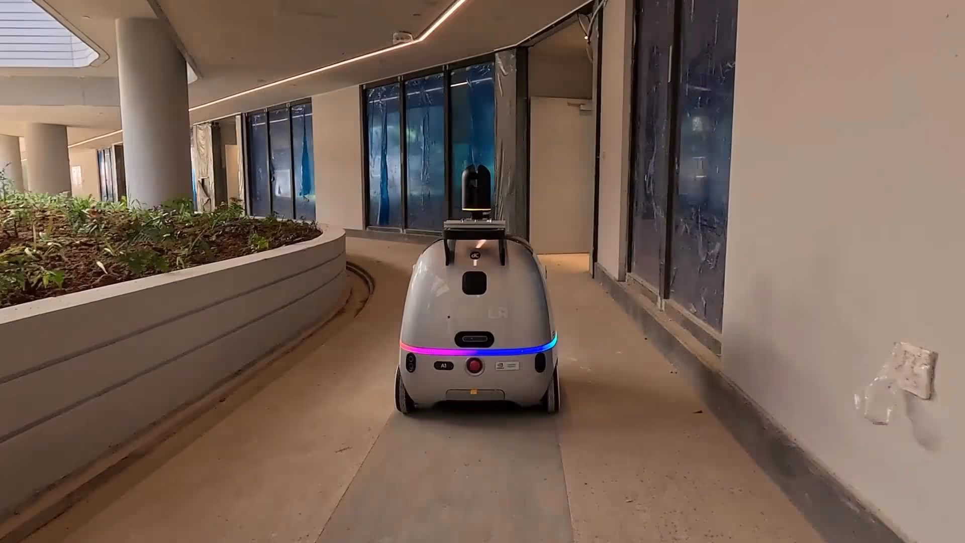 Small autonomous delivery robot with color LED lights moving along a corridor beside a planter.