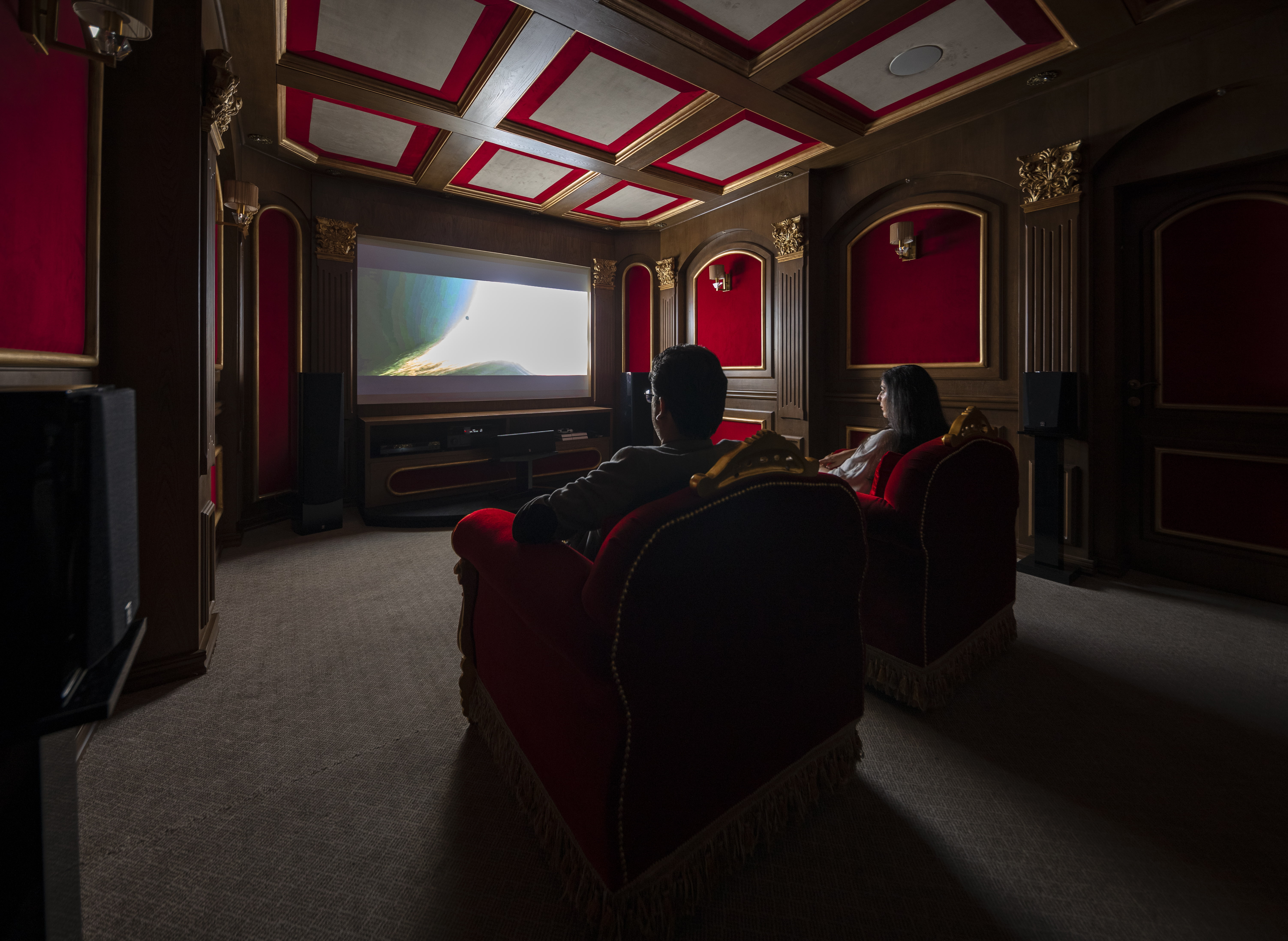 دو نفر در اتاق تئاتر خانگی با مبلمان قرمز روبه‌روی صفحه نمایش بزرگ مشغول تماشای فیلم هستند.
