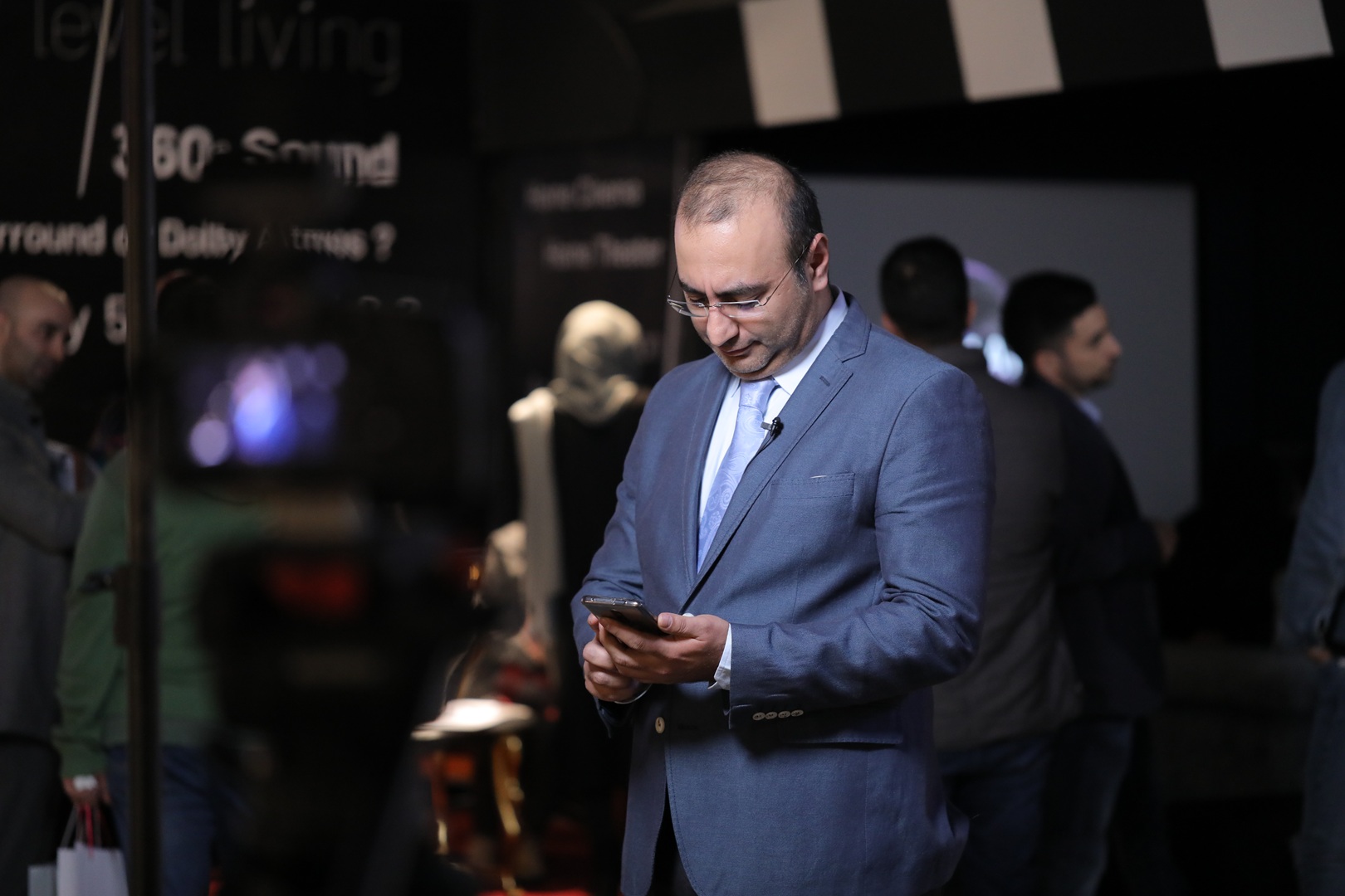 مردی با کت و شلوار آبی و کراوات روشن که به تلفن همراه خود نگاه میکند و در یک رویداد با افراد دیگر حضور دارد.