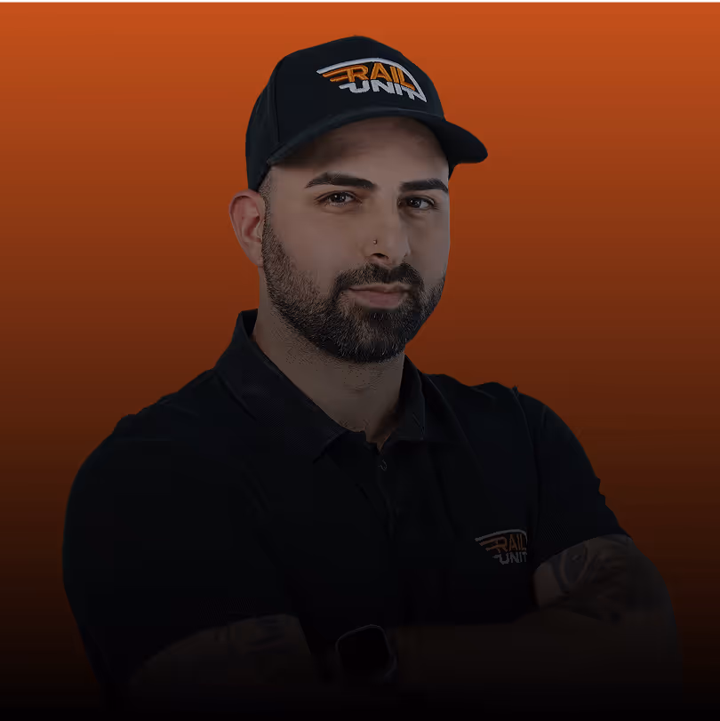 Mann mit Bart und Kurzhaarschnitt vor orangefarbenem Hintergrund, Blick nachdenklich, trägt schwarzes T-Shirt.