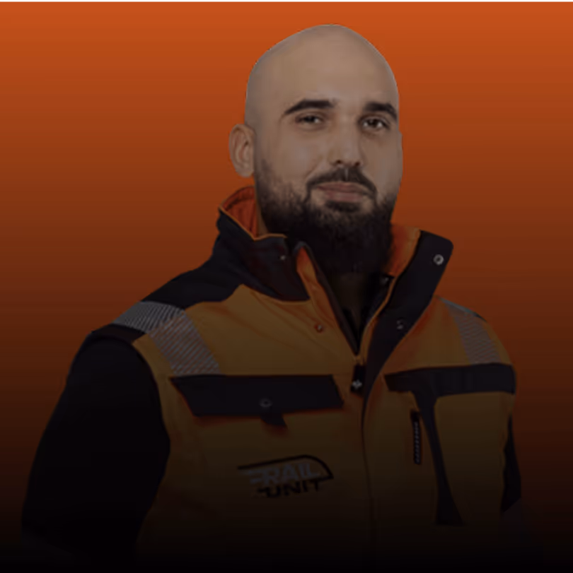 Mann in orangefarbener Arbeitsjacke vor rotem Hintergrund, schwarze Abzeichen, ernstes Gesicht.