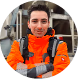 Mann in oranger Sicherheitsjacke vor Baustellengerät, lächelnd mit verschränkten Armen.