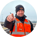 Mann in reflektierender Sicherheitsweste zeigt auf Kamera, steht auf Bahngleisen.