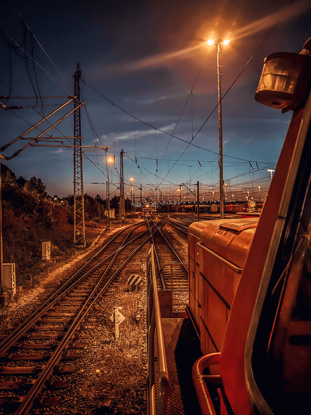 Nachtszenerie Zug fährt auf beleuchtetem Gleis unter blauem Himmel mit Oberleitungen und Laternen.