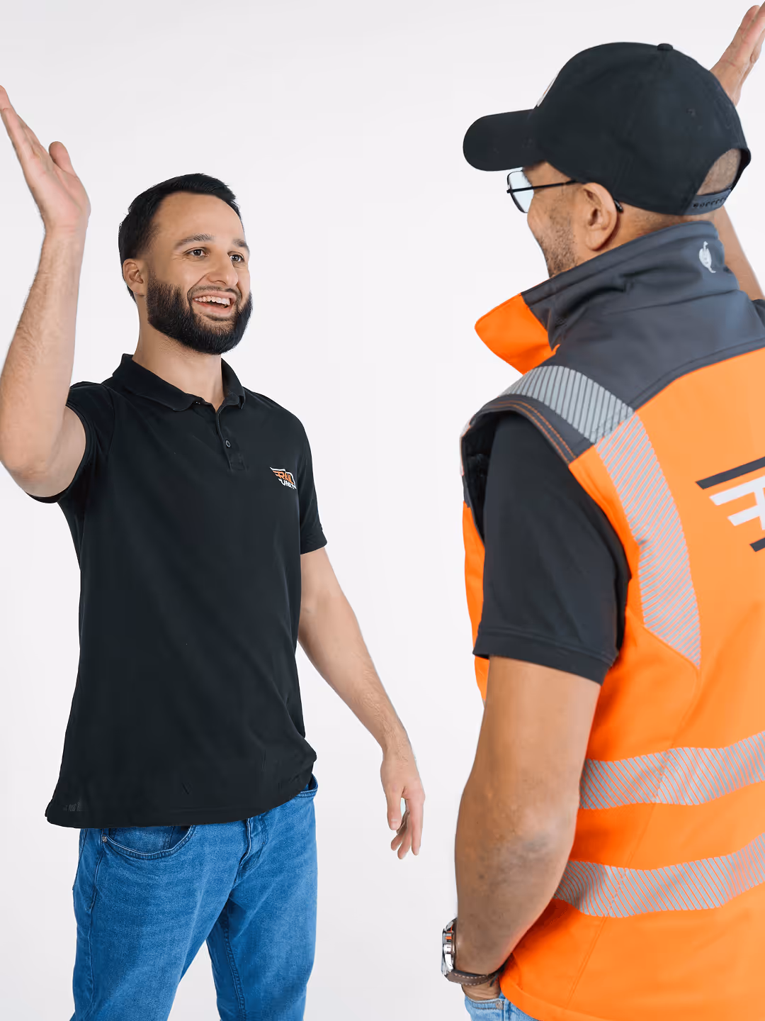 Zwei Männer im Gespräch, einer in schwarzem Polo, der andere in leuchtend orangefarbener Weste; entspannt und lächelnd.