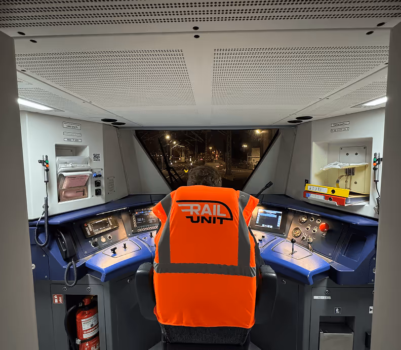 Mensch in roter Sicherheitsweste im modernen Zugcockpit, nachts; Fokus auf Bedienelemente und Armaturenbrett.