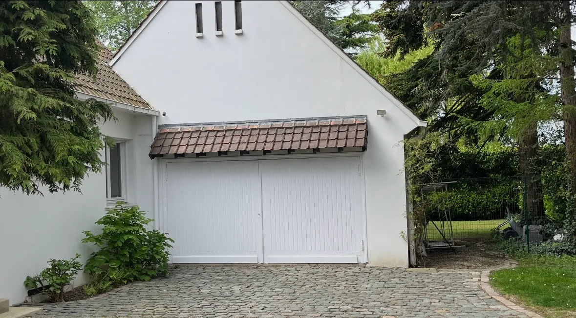 Maison blanche avec double porte de garage blanche et allée pavée entourée d'arbres verts.