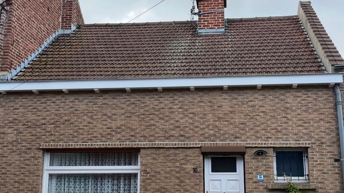 Façade de maison en brique avec toit en tuiles, cheminée, porte blanche et fenêtres avec grilles.