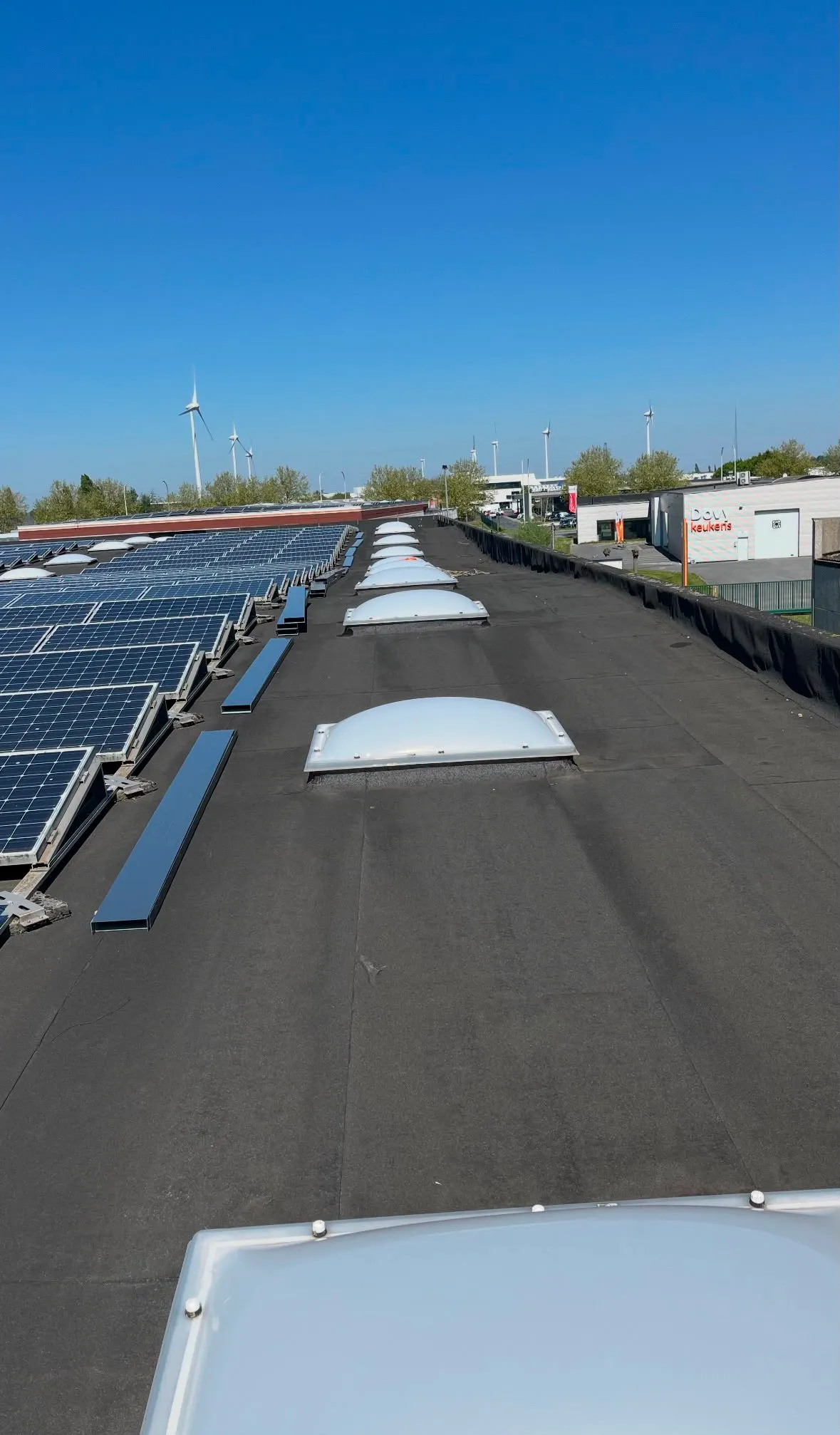 Toit plat avec panneaux solaires à gauche et éoliennes en arrière-plan sous un ciel bleu clair.