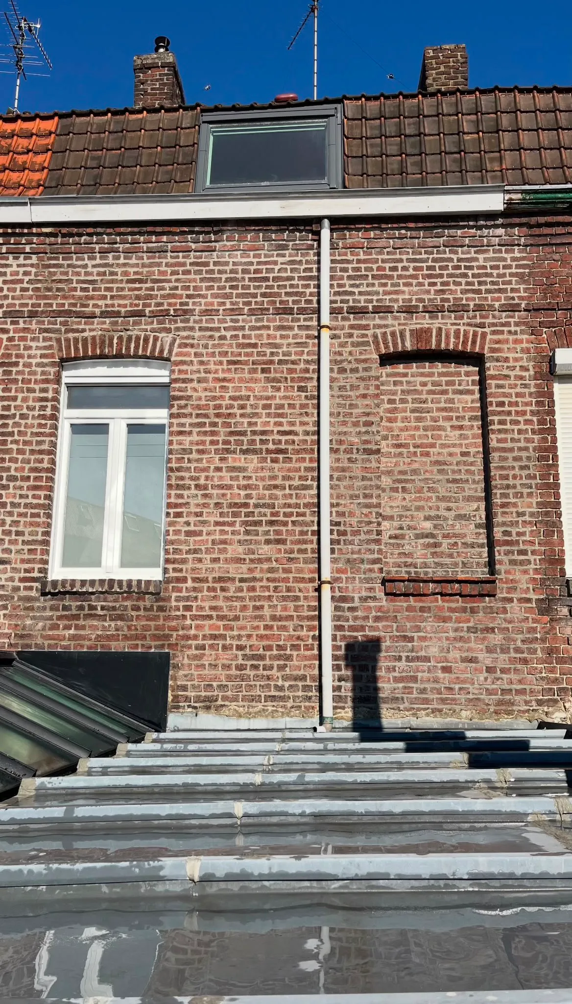 Façade en briques avec une fenêtre blanche, une fenêtre murée et une gouttière verticale sous un ciel bleu.
