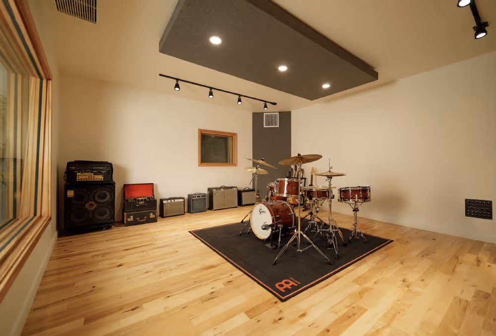 Modern music studio room with a drum set on a black rug and several guitar amplifiers along the wall.
