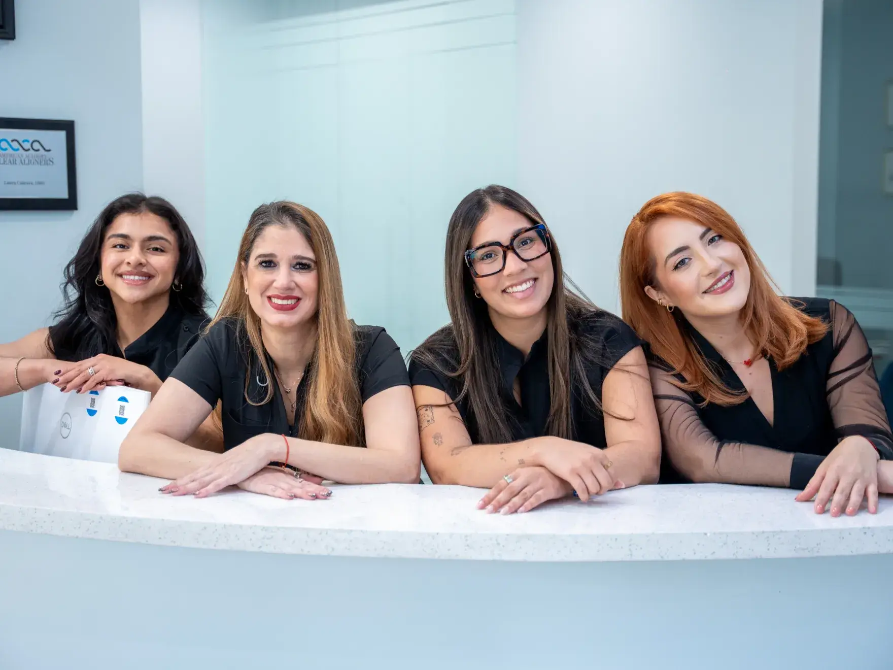 Four women in black shirts smiling and leaning on a white counter in an office setting.