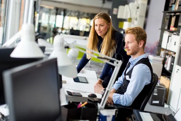 Two coworkers at a desk in the office: a woman leans over a document, a man works at a computer.