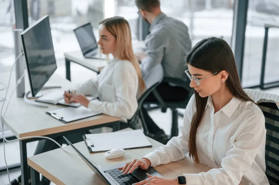 Bright office with large windows — two women and a man at computers, focused on work.