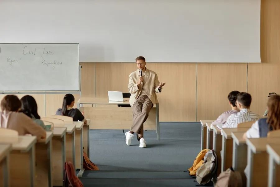 The lecturer conducts classes in the lecture hall; students sit in benches, next to a blackboard.