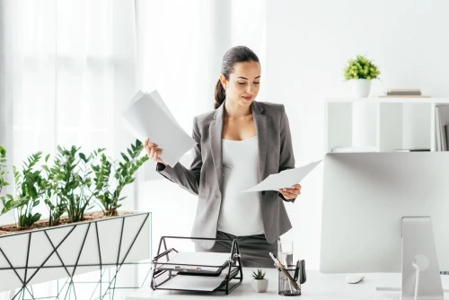 A woman in a bright office looks through documents while standing at a desk with a computer; plants in the background.