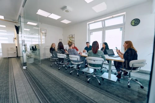 Modern office with glazed hall; a group of employees discuss the project at a common table.