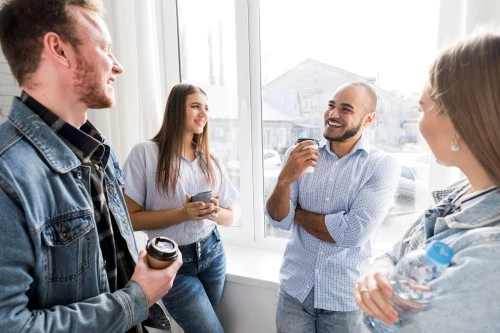 Four coworkers talk by the window during a break; smiles, cups of coffee in their hands.