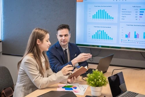 Meeting in the boardroom: a man in a suit discusses results on a chart screen, a woman takes notes; laptops and reports on a table.