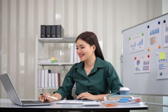 A woman in the office works at a laptop; on the board next to it hang printouts with graphs, notes and a mug on the desk.