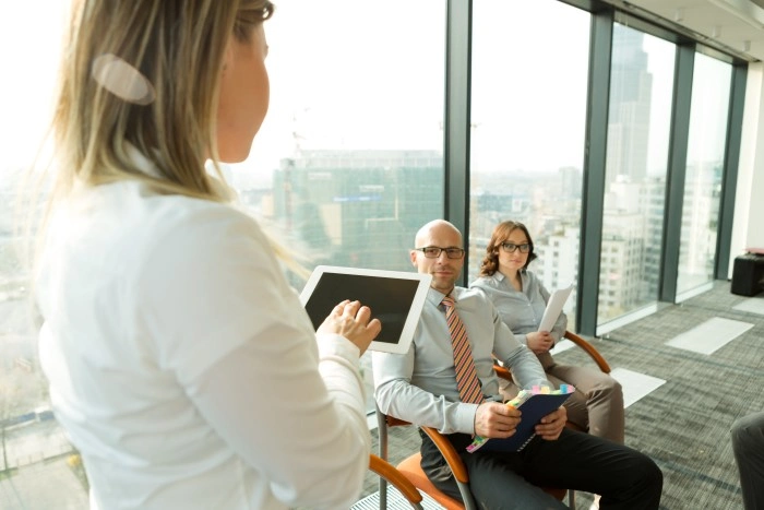 Presentation in a room with panoramic windows: a woman holds a tablet, two participants sit with notes; in the background a panorama of the city.