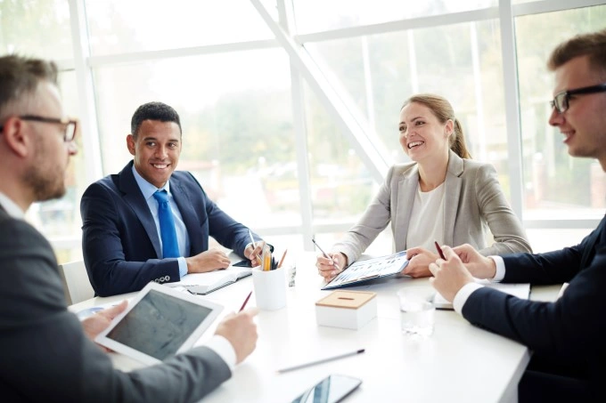 Meeting in a bright conference room: four people at the table discuss, one holds a tablet, documents and glasses of water on the countertop.