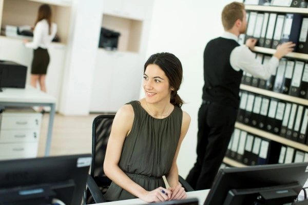 Modern workstation - monitors on the desk, the employee looks to the side; behind her shelves with documents.