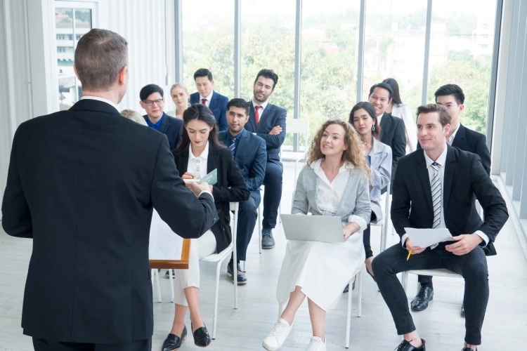 A rear-facing speaker conducts training; a group of listeners in business attire sits in a bright hall with large windows.